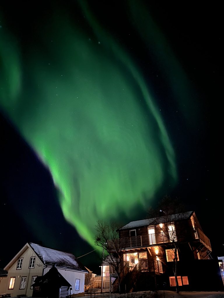 Des aurores boréales vertes illuminent le ciel nocturne au-dessus de l'écolodge Båthuset aux fenêtres éclairées à Sommaroy, près de Tromso - un voyage Norvège parfait pour ceux qui recherchent des vacances dans la nature et le respect de l'environnement.